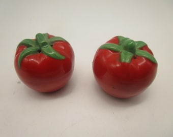Könnte beinhalten: Ein Paar Salz- und Pfefferstreuer aus Keramik in Form von runden roten Tomaten. Jeder Streuer hat einen glänzend roten Körper mit einem geformten grünen Stiel und Blättern an der Oberseite sowie kleinen Löchern zum Dosieren des Gewürzes.
