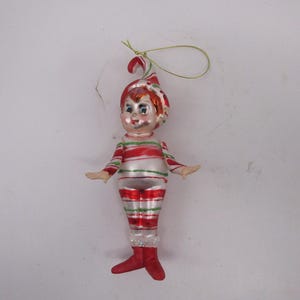 May include: A glass Christmas ornament shaped like an elf with a pointed hat, wearing a white suit with horizontal red and green stripes. The elf has a painted face with rosy cheeks, red lips, and red boots. A gold hanging loop is attached to the top.