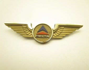 Könnte beinhalten: Eine goldfarbene Metallnadel in Form von Flügeln mit einem zentralen kreisförmigen Emblem. Das Emblem zeigt ein blaues Dreieck über einem roten horizontalen Balken und das Wort DELTA in weißen Großbuchstaben auf strukturiertem Goldgrund.