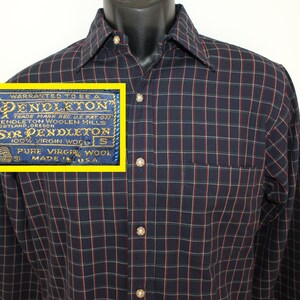 Pode incluir: Uma camisa de manga comprida azul e branca xadrez com uma faixa vermelha. A camisa tem colarinho de botões e um bolso no peito esquerdo. O rótulo diz "WARRANTED TO BE A PENDLETON" e "SIR PENDLETON 100% VIRGIN WOOL".