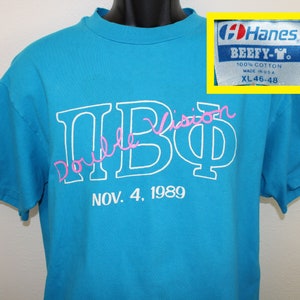 Könnte beinhalten: Ein türkisfarbenes T-Shirt mit einem weißen Hanes BEEFY-Y-Etikett. Das Shirt hat einen weißen Umriss der Buchstaben "ПВФ" mit dem Wort "Double Vision" in rosa Kursivschrift über den Buchstaben. Der Text "NOV. 4, 1989" ist unter den Buchstaben gedruckt.