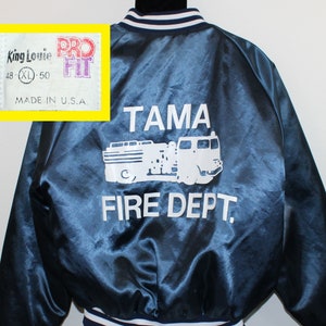 以下が含まれることがあります： 白い刺繍の消防車と「Tama Fire Dept.」の文字が背中に施された青いサテン製ボンバージャケット。ジャケットは白と青のストライプの襟と袖口が付いています。前面のラベルには「King Louie Pro Fit 48 XL 50 Made in U.S.A.」と記載されています。
