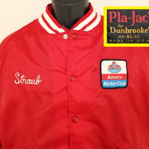 May include: Red satin bomber jacket with white stripes on the collar and cuffs. The jacket has a snap closure and features an embroidered "Straub" on the left chest and a "Pla-Jac by Dunbrooke® 48-XL-50 MADE IN USA" label on the right chest. The jacket also has an embroidered Amoco Motor Club patch on the right side.