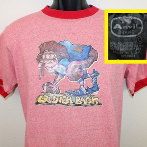 Puede incluir: Camiseta roja y gris con un jugador de fútbol de dibujos animados que lleva un casco y sostiene un balón de fútbol. La camiseta tiene un ribete rojo y el texto "ERDICH BACh" está impreso en la parte delantera. La etiqueta de la camiseta dice "Anvil Brand" e incluye el texto "50% Poliéster 50% Algodón Sin lejía Lavado a máquina en agua tibia - Secado por goteo Hecho en EE. UU. WPL10884".