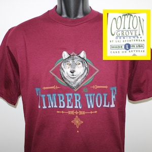 Puede incluir: Una camiseta de color granate con un gráfico de lobo y el texto "Timber Wolf" en azul y blanco. La camiseta está hecha de algodón y está hecha en los EE. UU. La etiqueta dice "Cotton Grove Designs by LSJ Sportswear Made in USA Care on Reverse."