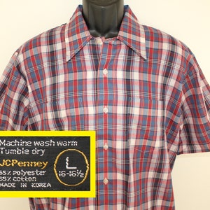 May include: A red, blue and white checked short-sleeved shirt with a button-down collar. The shirt has a pocket on the left chest and a label that reads "JCPenney 65% polyester 35% cotton MADE IN KOREA" and "Machine wash warm Tumble dry 16-16 1/2".