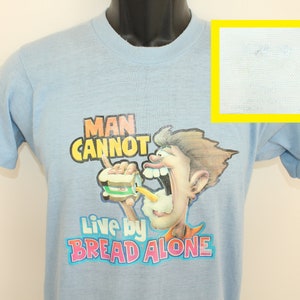 Puede incluir: Una camiseta azul claro con una ilustración de dibujos animados de una persona comiendo un sándwich. El texto en la camiseta dice "Man Cannot Live by Bread Alone."