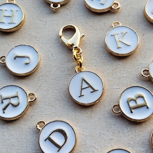 Könnte beinhalten: Goldfarbene Metallanhänger mit weißen Emaille-Mittelstücken, die jeweils einen anderen Buchstaben des Alphabets zeigen. Ein goldfarbener Verschluss ist an einem Anhänger befestigt. Die Anhänger sind rund mit einem goldenen Rand und einer kleinen Schlaufe zur Befestigung. Die Buchstaben sind in einer Serifenschrift.