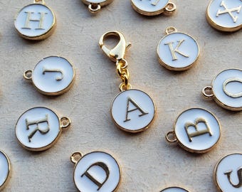 May include: Gold-coloured metal charms with white enamel centres, each displaying a different letter of the alphabet. A gold clasp is attached to one charm. The charms are round with a gold border and a small loop for attachment. The letters are in a serif font.