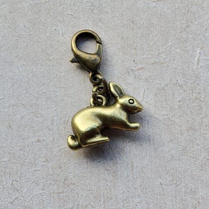 May include: A gold-coloured metal rabbit charm with a lobster clasp. The charm features a detailed rabbit design, including ears, eyes, and a tail. This charm is suitable for jewellery making or craft projects. The charm is made of metal.