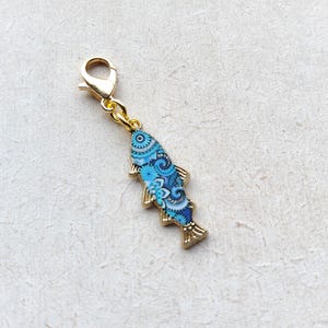 Könnte beinhalten: Ein goldfarbener Fisch-Anhänger mit einem Karabinerverschluss. Der Fisch ist mit einem blau-weißen Blumenmuster verziert. Der Anhänger ist etwa 3,8 cm lang.