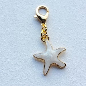 Könnte beinhalten: Ein goldfarbener Anhänger mit einem Karabinerverschluss und einer kleinen Kette. Der Anhänger zeigt einen weißen Seestern mit goldenen Verzierungen. Der Seestern hat die Form eines fünfzackigen Sterns. Der Anhänger ist wahrscheinlich für ein Armband oder eine Halskette.