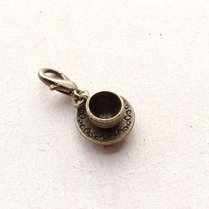 Può includere: Un ciondolo in bronzo con una tazza da tè e un piattino in miniatura. Il ciondolo ha un design decorativo e una chiusura a moschettone.