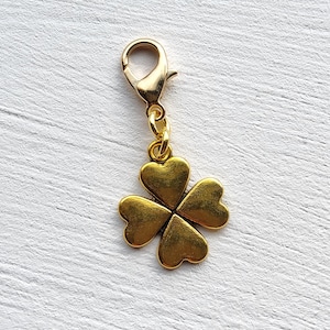 Peut inclure: Un charm doré avec un motif de trèfle à quatre feuilles. Le charm est attaché à un fermoir doré. Le trèfle a une surface texturée sur un fond blanc texturé.