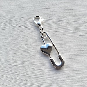 Op de afbeelding: Een zilverkleurige bedel met een veiligheidsspeldontwerp en een hartvormige hanger. De bedel heeft een karabijnsluiting voor bevestiging aan een armband of ketting. De hartvormige hanger is glad en reflecterend.