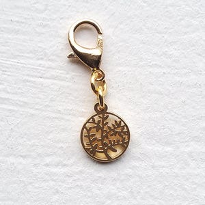 Könnte beinhalten: Ein goldfarbener Charm mit einem Baumdesign. Der Charm hat einen runden Anhänger mit einer ausgeschnittenen Baumsilhouette. Er ist mit einem Karabinerverschluss zur einfachen Befestigung an einem Armband oder einer Halskette versehen. Der Charm besteht aus glänzendem Metall.