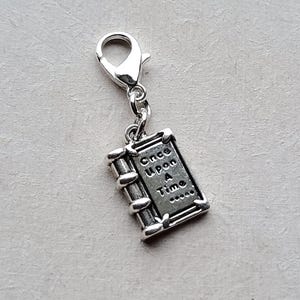 May include: Silver charm shaped like a book with the words "Once Upon A Time" engraved on the cover. The charm is attached to a silver lobster clasp.