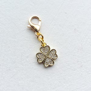 May include: A gold-toned metal charm featuring a four-leaf clover design encrusted with small, clear, sparkling stones. The clover is attached to a gold-toned lobster claw clasp by a small circular jump ring, creating a functional jewelry accessory.