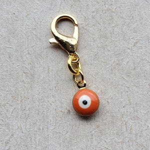 May include: A gold-colored lobster clasp with a small chain and a round orange charm. The charm features a white and black eye design, commonly known as an evil eye, for protection. This charm is a jewelry accessory.