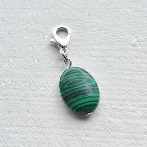May include: A silver-toned charm with a lobster clasp and a green malachite stone. The oval-shaped stone features dark green and black swirling patterns. This charm can be used for jewelry making or as a decorative accent.