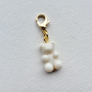 May include: A white gummy bear charm with a gold-coloured lobster clasp. The charm is a small, three-dimensional bear shape with rounded features. The clasp is attached to the bear's head with a small gold-coloured ring.