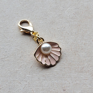 May include: A gold-colored charm with a light pink enameled seashell design, featuring a white pearl in the center. The charm has a lobster clasp for easy attachment. The seashell design is detailed and elegant.