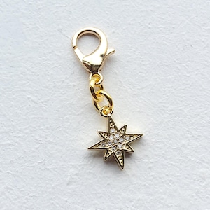 May include: A gold-coloured charm with a starburst design. The charm features a lobster clasp, a small chain, and a star pendant with embedded clear crystals. The star has eight points and is a decorative accessory.