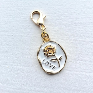 May include: Gold-coloured charm with a white enamel oval pendant featuring a gold rose and the word "LOVE". The charm has a lobster clasp for easy attachment to a bracelet or necklace.