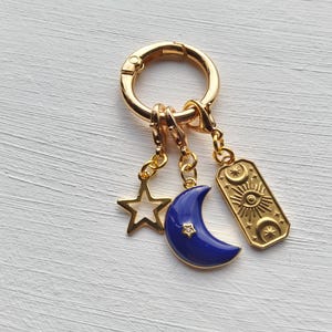 Puede incluir: Llavero dorado con colgantes celestiales. Incluye una estrella, una luna creciente azul y un dije rectangular con un diseño de ojo y sol. Los dijes cuelgan de un cierre dorado, creando un accesorio elegante.