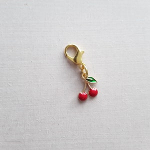 May include: Gold-tone charm with a red cherry and green leaf design. The charm has a lobster clasp.