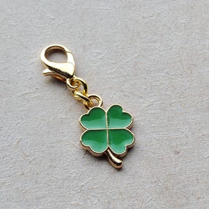 May include: A gold-coloured charm featuring a four-leaf clover design. The clover is green and has a gold-coloured stem.