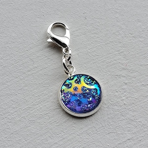 May include: A round charm with a silver-colored metal frame and clasp. The charm's center features a colorful, iridescent design with shades of blue, purple, and yellow. The charm is likely a pendant or accessory for jewelry or other crafts.