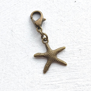 May include: A bronze-toned metal starfish charm attached to a matching lobster claw clasp. The starfish features a textured, bumpy surface detail on its front side, connected to the clasp by two small circular jump rings in a matching metal finish.