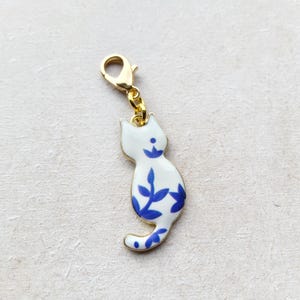 May include: A white cat charm with blue floral patterns and a gold-coloured clasp. The cat-shaped pendant is made of enamel and has a gold-coloured chain. The charm is a decorative accessory.