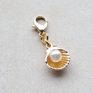 Könnte beinhalten: Goldfarbener Anhänger mit Muschel-Design und einer Perle im Inneren. Der Anhänger hat einen Karabinerverschluss.
