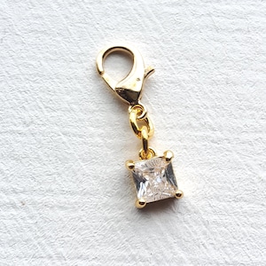 May include: A gold-coloured charm featuring a square, clear gemstone. The charm has a lobster clasp and a small chain connecting to the gemstone. The gemstone is set in a gold-coloured frame with four small prongs.