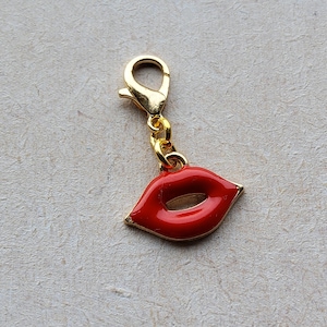 May include: A gold-colored charm with a lobster clasp and a red lips pendant. The lips are glossy red with a gold outline. The charm is likely for a bracelet or necklace.