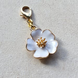 Puede incluir: Un charm de flor blanca con detalles dorados. La flor tiene un centro dorado y un contorno dorado. El charm está unido a un cierre dorado.