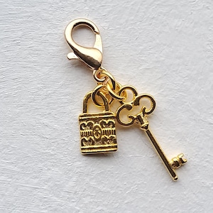Puede incluir: Charm dorado con diseño de candado y llave. El candado tiene un patrón ornamentado y la llave tiene una cabeza detallada. El charm está unido a un cierre dorado, adecuado para joyería o proyectos de artesanía.