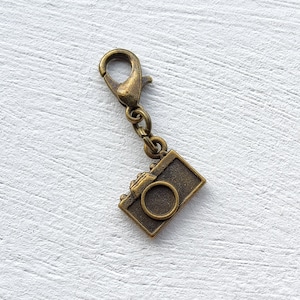 May include: A bronze-colored camera charm with a lobster clasp. The rectangular charm features a detailed camera design with a circular lens. The charm is attached to a small chain and is set against a white textured background.