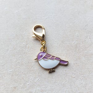 Op de afbeelding: Een goudkleurige vogelbedel met een karabijnsluiting. De vogel is wit met paarse accenten op zijn vleugels en staart. De bedel is waarschijnlijk gemaakt van metaal en is ontworpen om aan een armband of ketting te worden bevestigd.