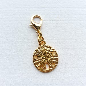 May include: A gold-coloured charm featuring a sand dollar design. The round charm has a detailed pattern and is attached to a lobster clasp. The charm is made of metal and is set against a white background.