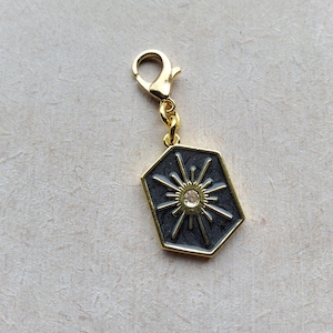 May include: Gold-coloured charm with a sunburst design. The charm has a dark grey background, a gold border, and a small, clear crystal in the centre. It includes a gold-coloured clasp for attachment.