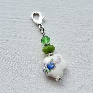 Pode incluir: Um berloque de prata com um pingente de flor de porcelana branca com um design floral azul. O berloque inclui contas verdes e um fecho prateado. Um acessório decorativo.