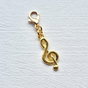 May include: A gold-colored treble clef charm with a lobster clasp. The musical note charm is suspended from a small chain and is displayed against a white, textured surface. The charm is a decorative accessory.