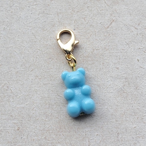 Könnte beinhalten: Ein hellblauer Gummibärchen-Anhänger mit einem goldfarbenen Karabinerverschluss. Der Anhänger ist eine Miniatur, dreidimensionale Darstellung eines Gummibärchens. Der Hintergrund ist eine strukturierte, cremefarbene Oberfläche.