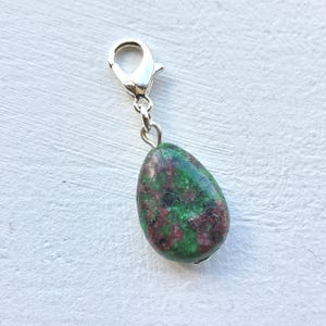 May include: A teardrop-shaped charm with a silver-colored clasp. The charm is a mix of green and red colors, with a speckled pattern. The charm is likely made of stone or a similar material. The clasp is attached to the top of the charm.