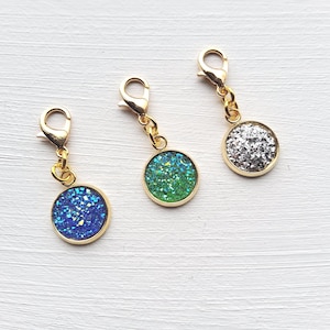 Op de afbeelding: Drie goudkleurige bedelhangers met ronde, met glitter gevulde centra in blauw, groen en zilver. Elke bedel heeft een karabijnsluiting voor eenvoudige bevestiging. De hangers worden getoond op een wit, getextureerd oppervlak.