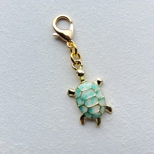 Könnte beinhalten: Ein goldfarbener Schildkröten-Anhänger mit Karabinerverschluss. Die Schildkröte hat ein hellblaues und weißes Schalendesign. Der Anhänger ist ein Schmuckzubehör.