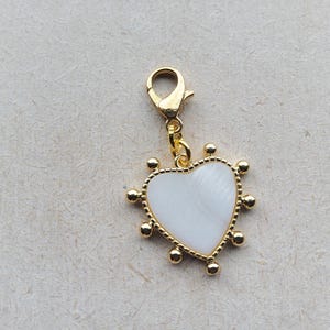 Può includere: Un ciondolo a forma di cuore color oro con un centro bianco e un moschettone. Il ciondolo è adornato con piccole sfere color oro attorno al perimetro del cuore. Il ciondolo è progettato per la creazione di gioielli.